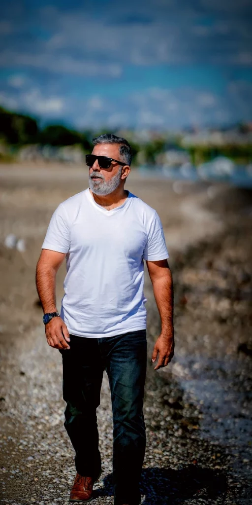 Graumelierter Mann trägt ein weißes T-Shirt und läuft entspannt am Strand entlang.