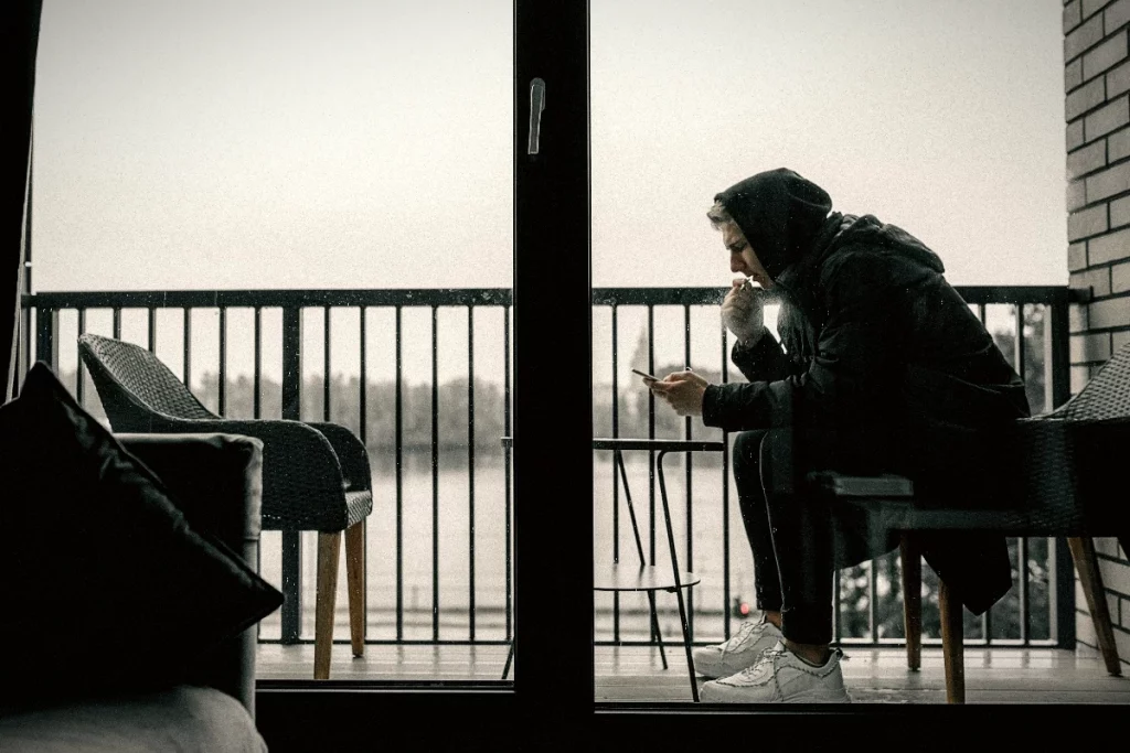 Mann sitzt allein auf einem Balkon, schaut auf sein Handy und wirkt einsam und angespannt.