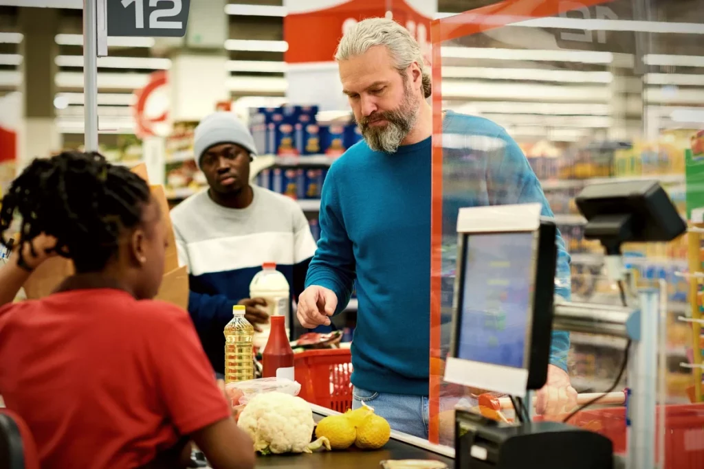 Mann mit grauem Bart steht an einer Supermarktkasse und wirkt in der Alltagssituation leicht verloren.