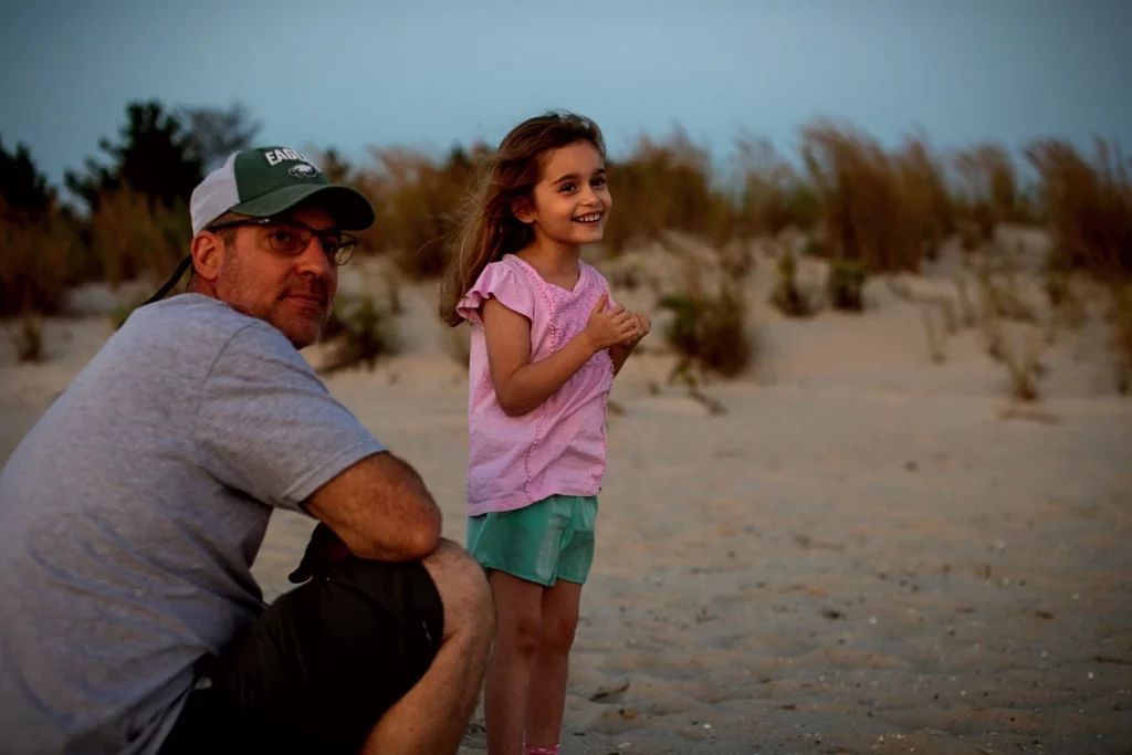 Vater mit Baseballkappe sitzt am Strand neben seiner Tochter und schaut nachdenklich.