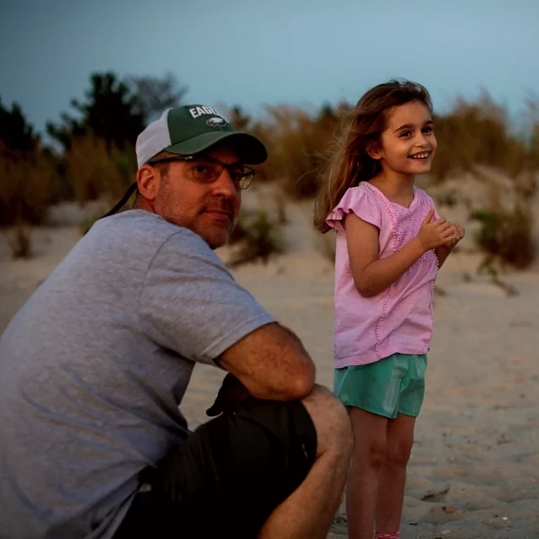 Vater mit Baseballkappe sitzt am Strand neben seiner Tochter und schaut nachdenklich.