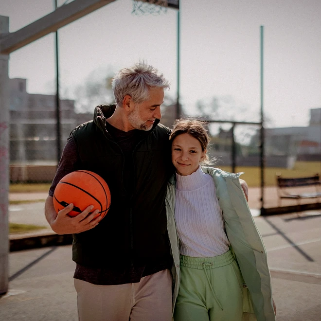 Vater läuft mit seiner Tochter über einen Basketballplatz und hält einen Ball unter dem Arm.