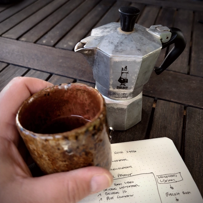 Eine Bialetti Moka-Express-Espressokanne steht auf einem Holztisch im Freien, davor hält eine Hand eine handgemachte braune Keramiktasse, im Vordergrund liegt ein aufgeschlagenes Notizbuch mit handschriftlichen Notizen.