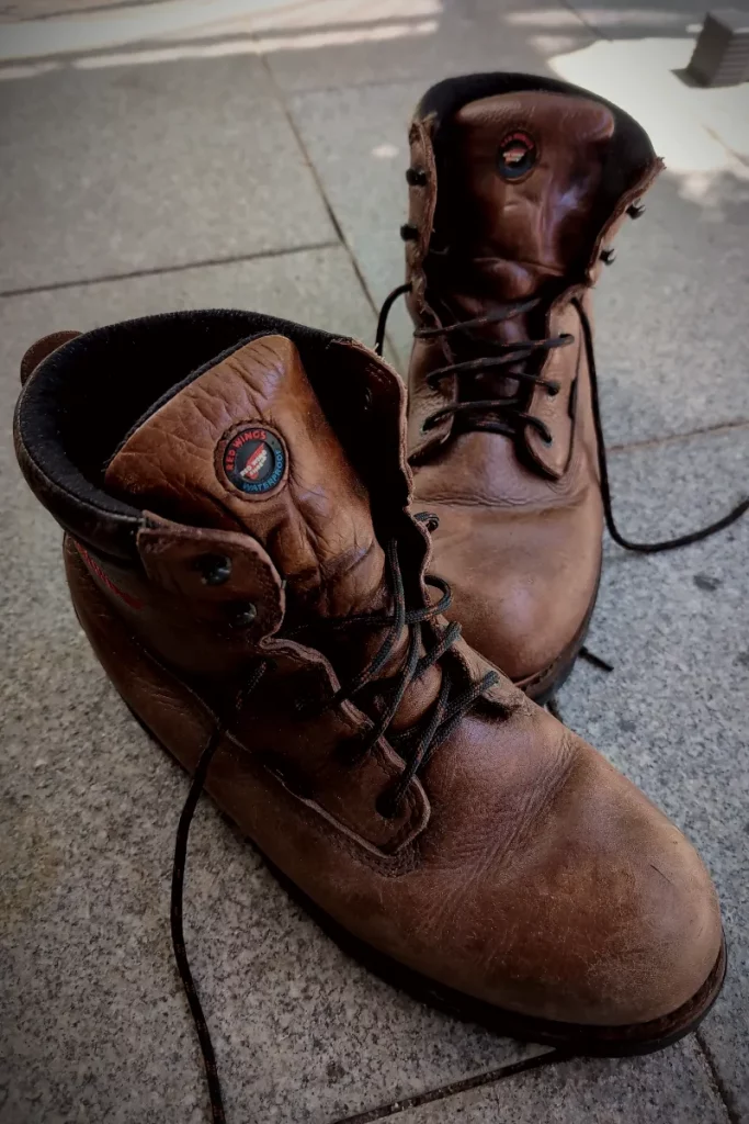 Ein Paar braune Red-Wing-Lederstiefel mit deutlichen Gebrauchsspuren steht auf grauem Pflaster, die Schnürsenkel teilweise gelöst.