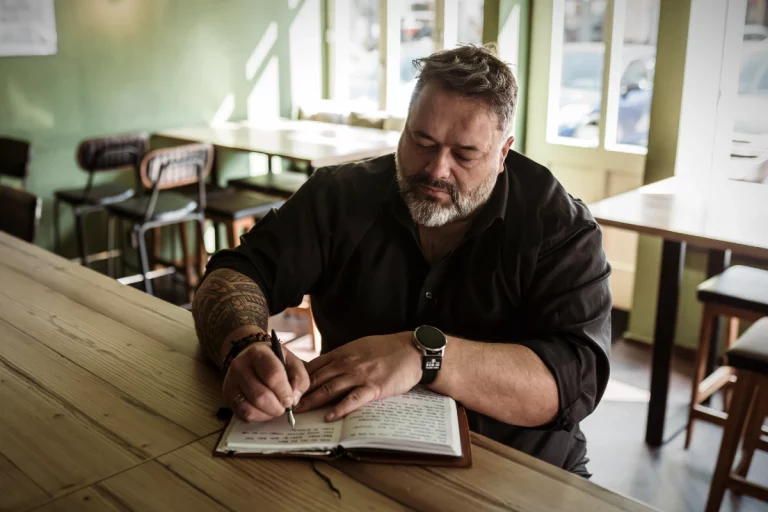 Mann mittleren Alters mit grauem Bart sitzt an einem Holztisch in einem Café und schreibt mit einem Füller in ein aufgeschlagenes Ledernotizbuch.