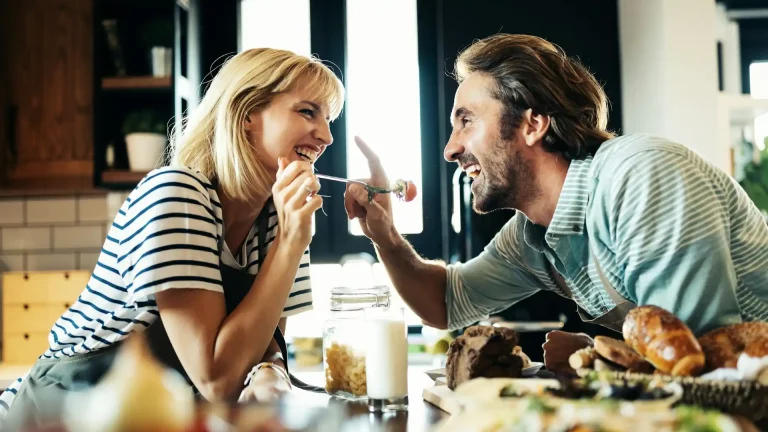 Paar mittleren Alters lacht sich beim gemeinsamen Essen in der Küche an.