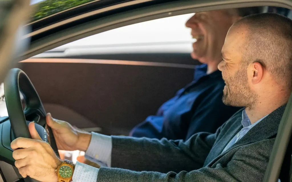 Zwei Männer sitzen gemeinsam im Auto; der Fahrer blickt während der Fahrt zu seinem Beifahrer.